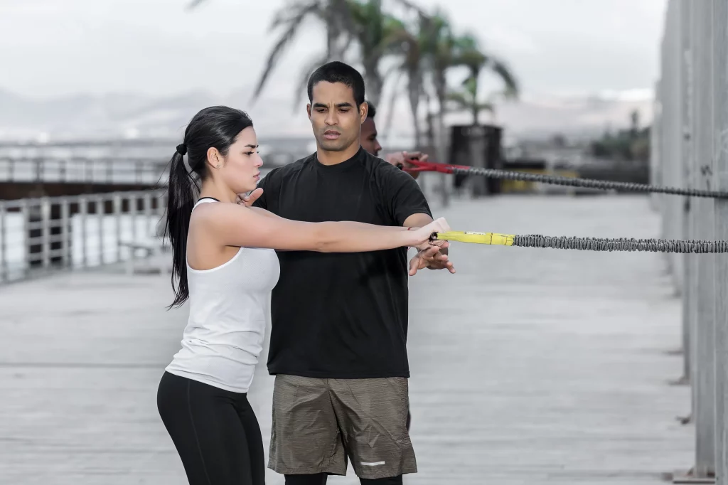 Adult sport instructor holding arms of attractive female and help her to perform exercise with resistance band properly during outdoor workout